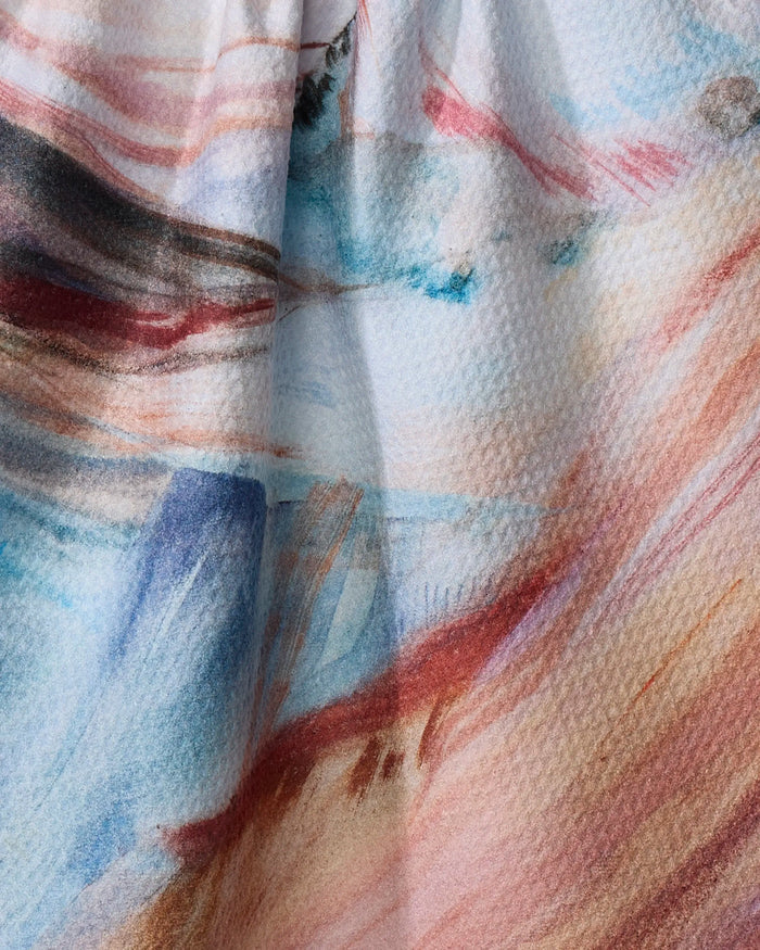 Close-up of Kelly Peters' "Golden Hour in the Rockies" tea towel, made from recycled materials, with soft watercolor-like streaks in blue, pink, brown, and red tones, gentle folds, and subtle shadows.