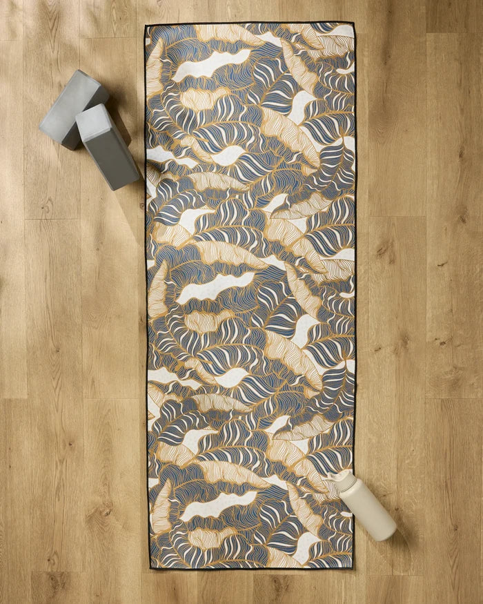 The Golden Palms yoga mat by Marina Solodka, featuring abstract blue, gold, and white designs, lies on a wooden floor. A non-slip towel covers the mat with two gray yoga blocks and a light water bottle placed beside it.