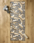 The Golden Palms yoga mat by Marina Solodka, featuring abstract blue, gold, and white designs, lies on a wooden floor. A non-slip towel covers the mat with two gray yoga blocks and a light water bottle placed beside it.