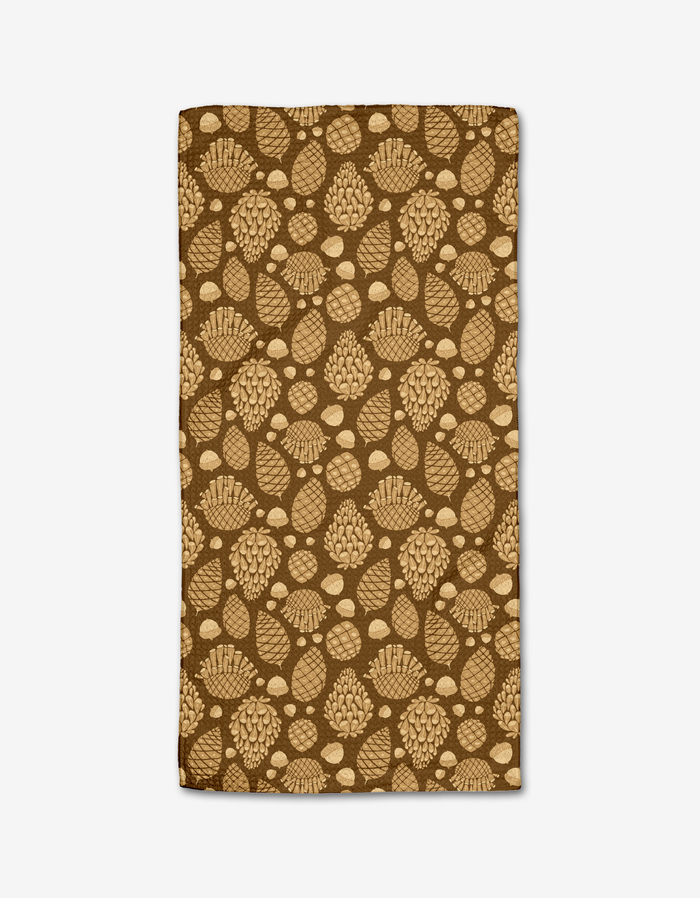 The Golden Pines Bar Towel by Danelys Sidron is a rectangular brown towel made from recycled materials, featuring a repeating pattern of tan pine cones and acorns on a white background.