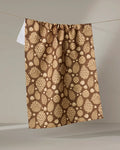 The "Golden Pines" tea towel by Danelys Sidron, featuring light beige pinecones and leaves on a brown background, hangs from a string against a neutral backdrop.