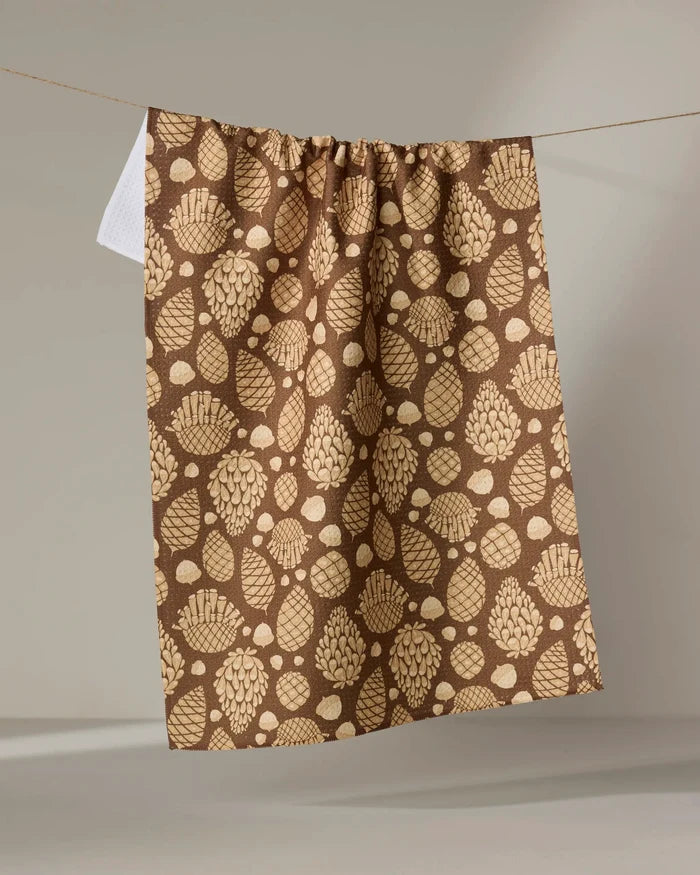 The "Golden Pines" tea towel by Danelys Sidron, featuring light beige pinecones and leaves on a brown background, hangs from a string against a neutral backdrop.