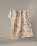 The "Golden Poppy" tea towel by Julianne Haness, featuring yellow and teal floral patterns and made from recycled materials, hangs over a clothesline against a neutral background.
