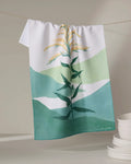 The Goldenrod tea towel by Elana Gabrielle, made from recycled materials and showcasing a green and yellow plant design, hangs on a line with stacked white bowls in the lower right corner against a neutral background.