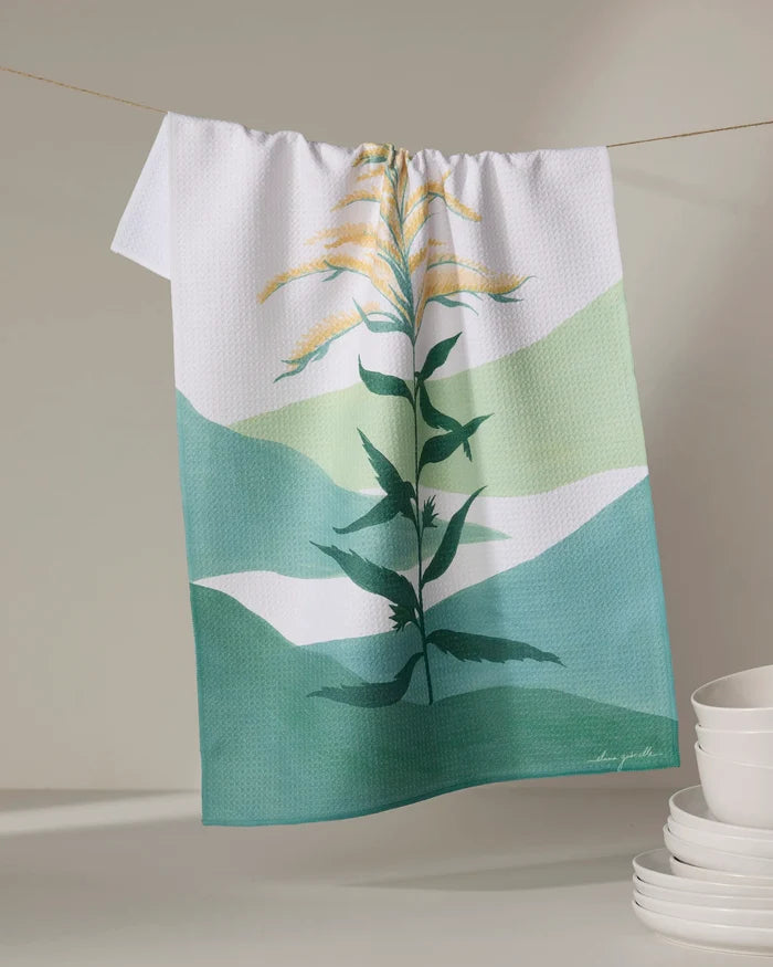 The Goldenrod tea towel by Elana Gabrielle, made from recycled materials and showcasing a green and yellow plant design, hangs on a line with stacked white bowls in the lower right corner against a neutral background.
