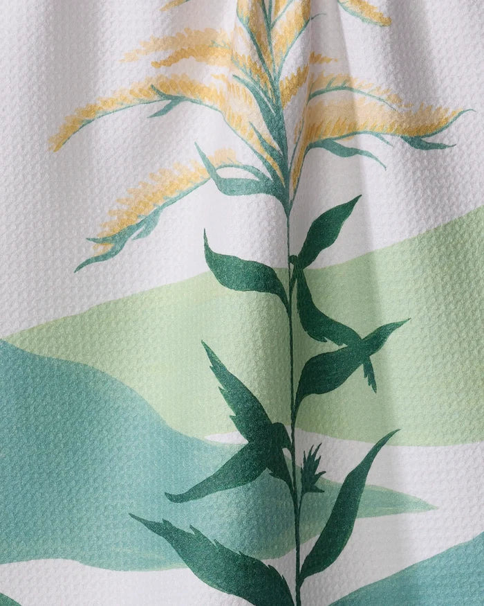 A close-up of the Elana Gabrielle Goldenrod kitchen tea towel, featuring a green and yellow botanical print with leaves and flower spikes on a white and light green background, made from recycled materials.