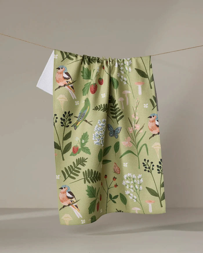 The "Good Green Earth" recycled kitchen towel by Janna Sue Design, with a botanical and bird print of flowers, leaves, butterflies, and finches, hangs on a string against a neutral background.