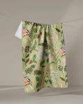 The "Good Green Earth" recycled kitchen towel by Janna Sue Design, with a botanical and bird print of flowers, leaves, butterflies, and finches, hangs on a string against a neutral background.