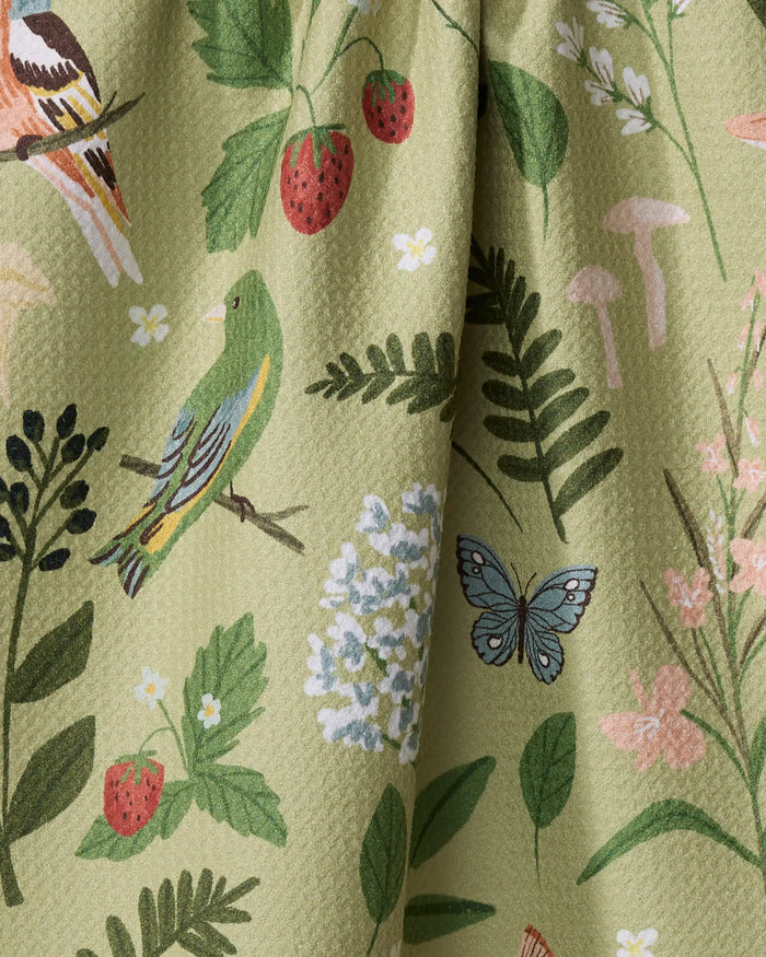 A close-up of Janna Sue Design’s Good Green Earth recycled kitchen towel, featuring a nature-themed print with birds, strawberries, leaves, flowers, mushrooms, and a blue butterfly on a light green textured background.