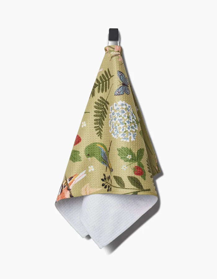 The Good Green Earth bar towel by Janna Sue Design features a floral, butterfly, and bird pattern in green. Made from recycled materials, it hangs from a black loop and has a white textured inner side.