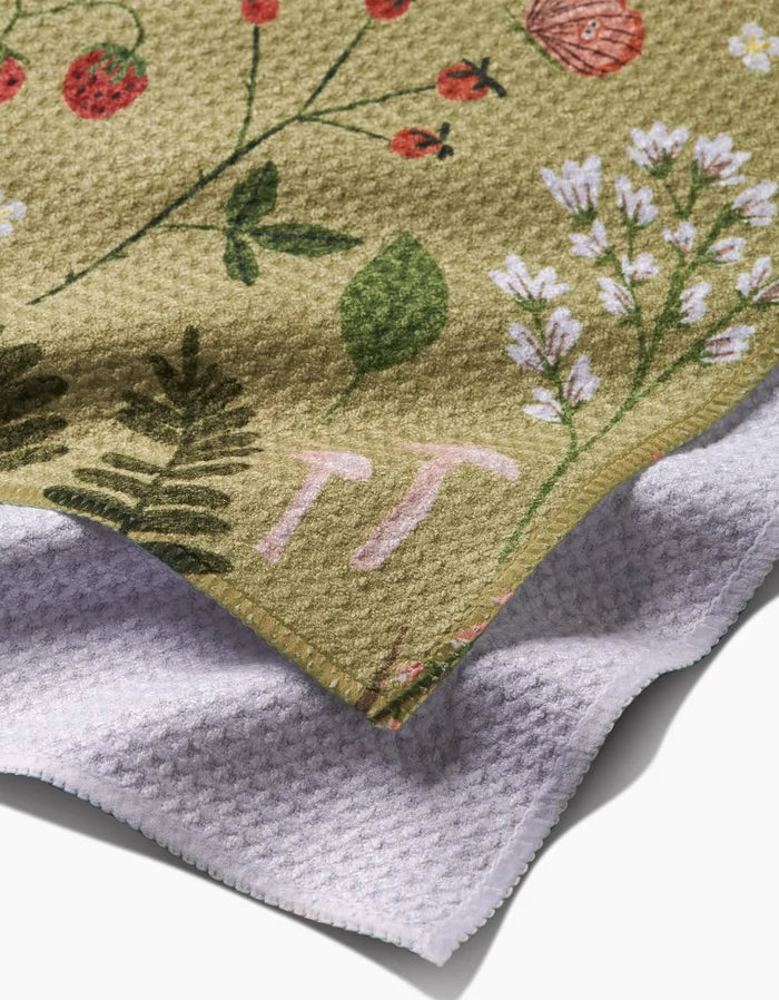 A close-up of the Good Green Earth bar towel by Janna Sue Design, featuring a botanical print on Geoweave fabric with embroidered pink TT initials and a soft white underside, set against a tan background.