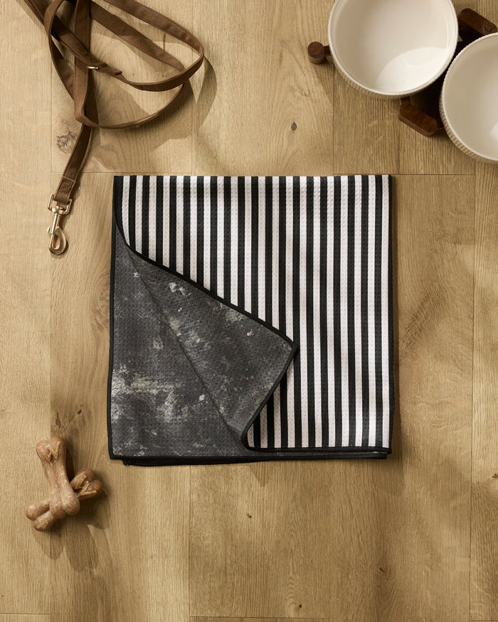 The Geometry Good Pup towel, a black and white striped, highly absorbent dog towel, is neatly folded on a wooden floor next to two white bowls, a brown leash, and bone-shaped dog toys.