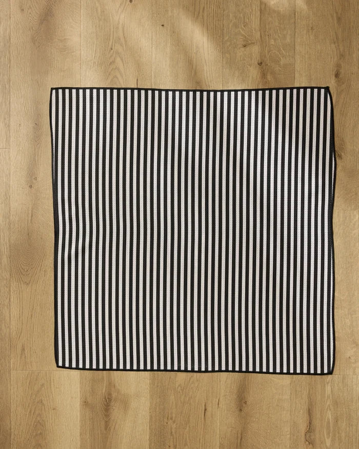 The Geometry Good Pup mat, a rectangular and highly absorbent piece with black and white vertical stripes, rests on a light wooden floor illuminated by natural light.