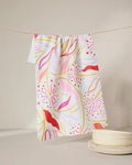 The GEOMETRY Grand Bloom kitchen towel, made from recycled materials, features vibrant pink, red, yellow, and orange patterns. Hanging on a line beside white textured plates on a softly lit surface, it brings color to any space.
