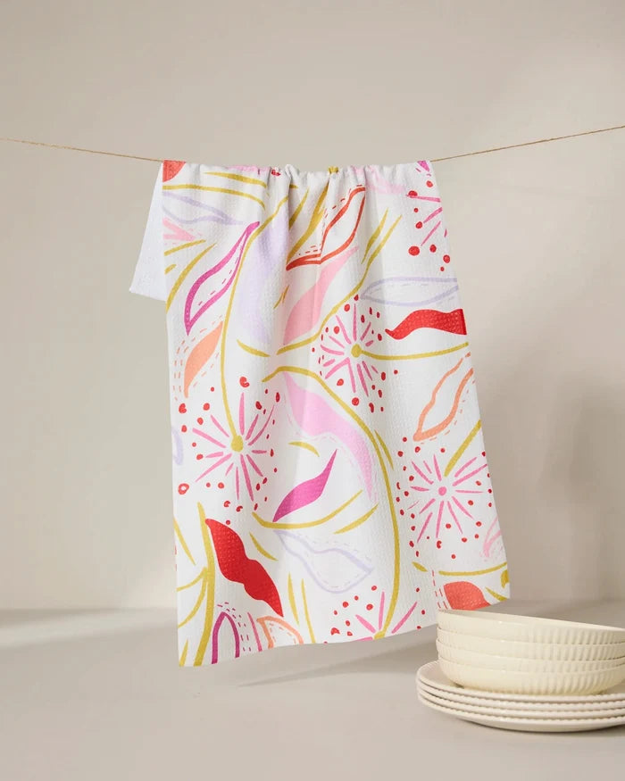 The GEOMETRY Grand Bloom kitchen towel, made from recycled materials, features vibrant pink, red, yellow, and orange patterns. Hanging on a line beside white textured plates on a softly lit surface, it brings color to any space.
