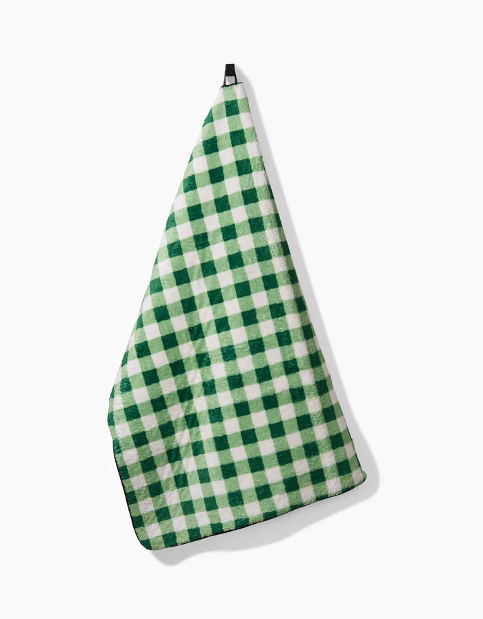The Grass Plaid dish towel by Danelys Sidron, featuring a green and white checkered design, hangs on a hook against a white background.
