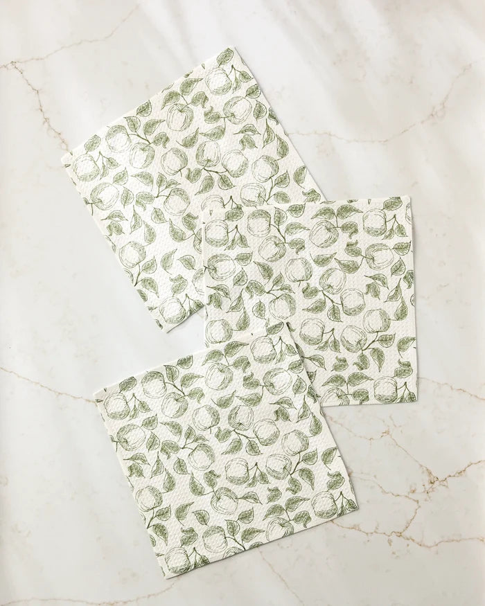 Three Gravenstein Apple napkins by Ash Cascade, made from recycled materials, feature a green leafy fruit pattern and are arranged with overlap on a light marble surface.
