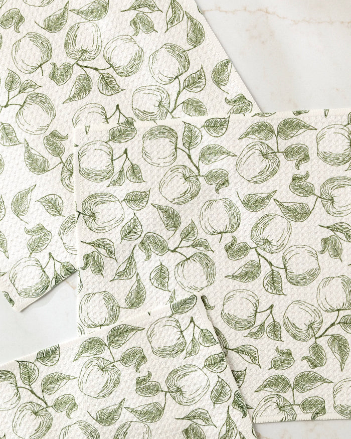 Ash Cascade's super-absorbent Gravenstein Apple dishtowel set features green line drawings of apples and leaves on white cloths, elegantly arranged on a light marble surface.