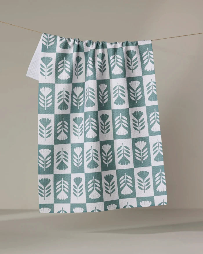 The Green Flower Patch tea towel by Charlie Rowan Designs, featuring a green and white geometric leaf pattern, hangs on a clothesline against a plain beige background.