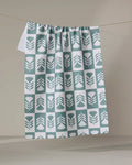The Green Flower Patch tea towel by Charlie Rowan Designs, featuring a green and white geometric leaf pattern, hangs on a clothesline against a plain beige background.