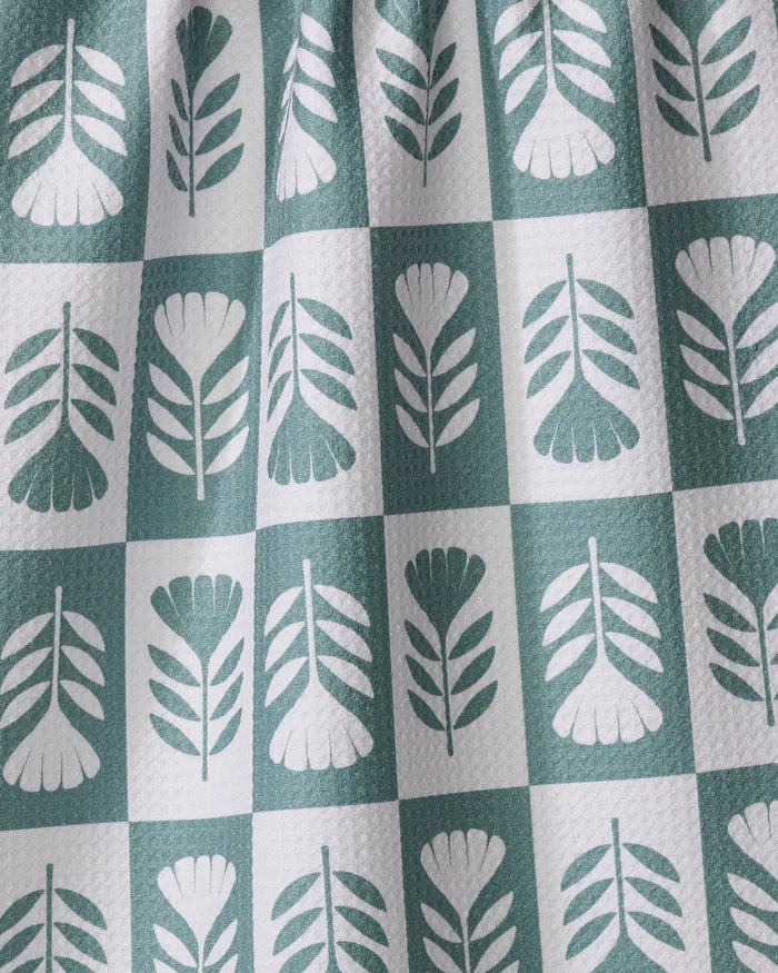 The Green Flower Patch tea towel by Charlie Rowan Designs features a green and white checkerboard pattern with stylized leaf motifs. Its textured, ultra-absorbent fabric and subtle folds add visual appeal.