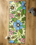 The GEOMETRY Green Flowers yoga mat, showcasing blue, white, and pink floral patterns with green leaves, lies on a wooden floor. Nearby are a non-slip yoga towel, a rolled white towel, and two gray yoga blocks.