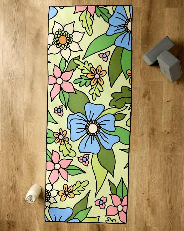 The GEOMETRY Green Flowers yoga mat, showcasing blue, white, and pink floral patterns with green leaves, lies on a wooden floor. Nearby are a non-slip yoga towel, a rolled white towel, and two gray yoga blocks.