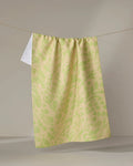 The Green Spring tea towel by Sareka Unique, featuring a yellow and light green pattern and crafted from recycled materials, hangs on a clothesline with a small white section at the top left against a neutral backdrop.