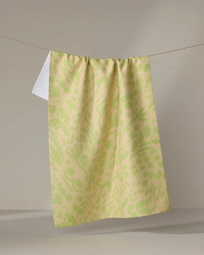 The Green Spring tea towel by Sareka Unique, featuring a yellow and light green pattern and crafted from recycled materials, hangs on a clothesline with a small white section at the top left against a neutral backdrop.