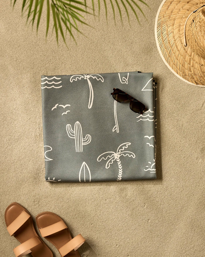 A flat lay of a beach scene on sand featuring Hufton Studio's Green Summer Vibes recycled blanket, sunglasses, a straw hat, brown sandals, and a palm leaf casting a shadow.