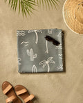 A flat lay of a beach scene on sand featuring Hufton Studio's Green Summer Vibes recycled blanket, sunglasses, a straw hat, brown sandals, and a palm leaf casting a shadow.