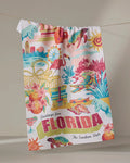 The Geometry "Greetings From Florida" tea towel, crafted from recycled materials, showcases vibrant Florida motifs—flamingos, turtles, citrus, beaches, hotels—and bold “Greetings from FLORIDA The Sunshine State” lettering.