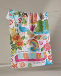 The Geometry "Greetings From Hawaii" tea towel features ultra-absorbent fabric and vibrant Hawaiian-themed designs—flowers, hula dancer, ukulele, and more—with "Greetings from HAWAII The Aloha State" text. Shown hanging on a line.