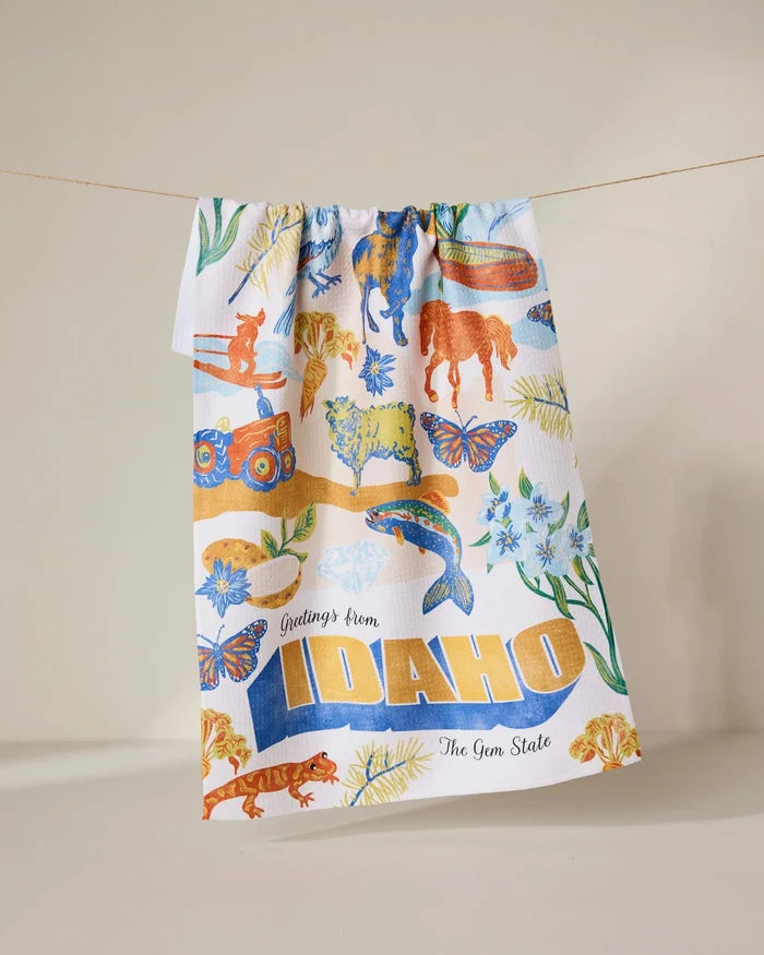 The Geometry Greetings From Idaho Kitchen Tea Towel hangs on a line, showcasing Idaho-themed wildlife, flowers, and scenery in colorful Geoweave fabric with bold yellow text: Greetings from IDAHO The Gem State.