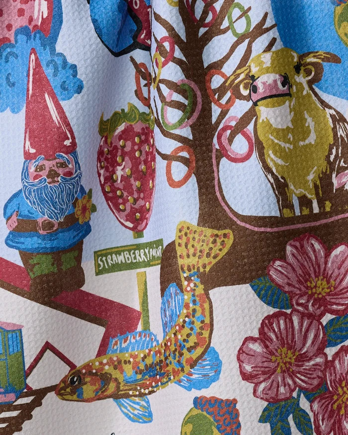 Geometry's "Greetings From Iowa" tea towel features a gnome, cow, strawberry, fish, flowers, tree, and a "Strawberry Point" street sign on a light background. Made from quick-drying recycled materials.