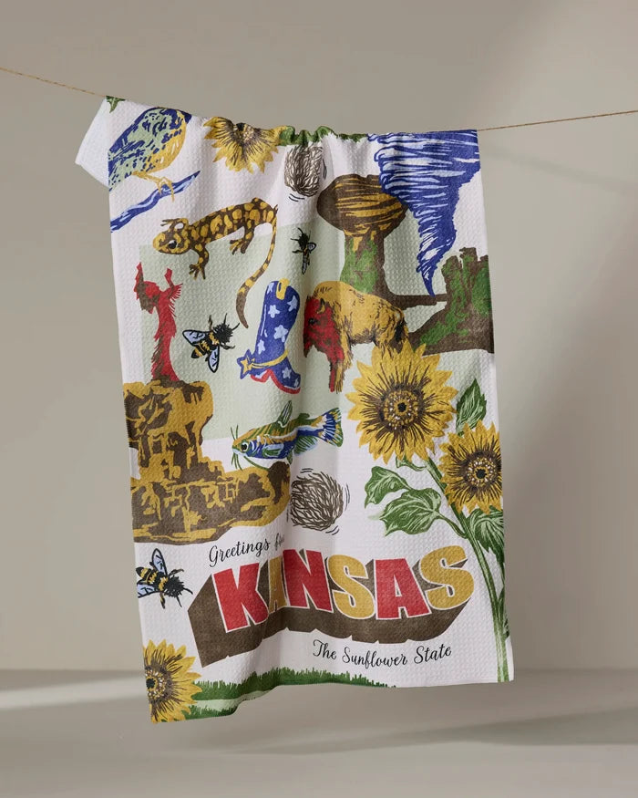 The Geometry "Greetings From Kansas" tea towel features colorful illustrations of Kansas landmarks, sunflowers, animals, and "Greetings from KANSAS The Sunflower State," adding charm and function to any kitchen. Ultra-absorbent design.