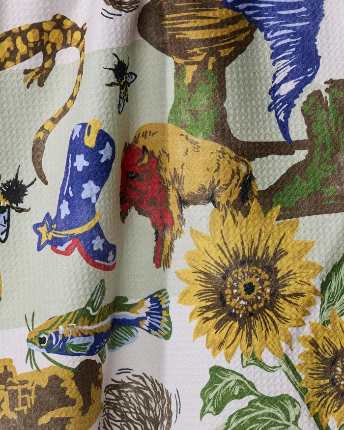 “Greetings From Kansas” by Geometry is a recycled kitchen towel with colorful illustrations—yellow sunflower, red and brown bison, blue cowboy boot, fish, insects, reptiles, and cacti—on a textured white background.