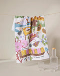 The Geometry Greetings From Montana tea towel, made from recycled materials, displays illustrated landmarks—flowers, trout, mountains, pine cones—with Greetings from Montana The Treasure State. Three glass containers accompany it.
