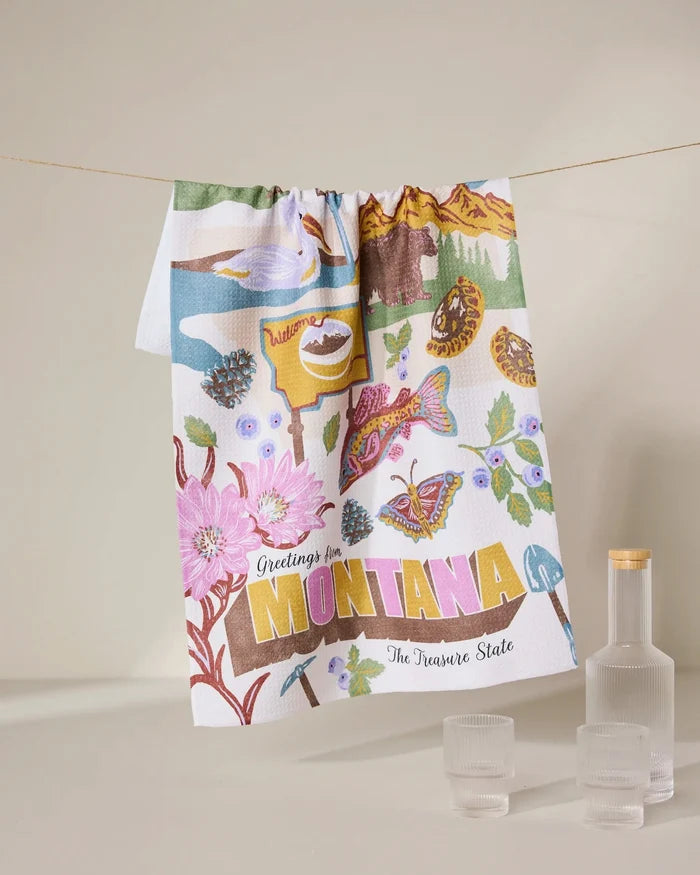 The Geometry Greetings From Montana tea towel, made from recycled materials, displays illustrated landmarks—flowers, trout, mountains, pine cones—with Greetings from Montana The Treasure State. Three glass containers accompany it.