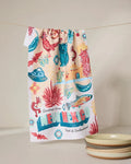 The Geometry Greetings From New Mexico dish towel hangs on a line, decorated with roadrunners, cacti, chili peppers, and the phrase “Greetings from New Mexico: Land of Enchantment.” Three stacked bowls are nearby.