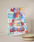 The Geometry Greetings From New York kitchen towel, made from recycled materials and featuring New York landmarks, roses, a blue cat, and text, hangs on a line beside three stacked plates.