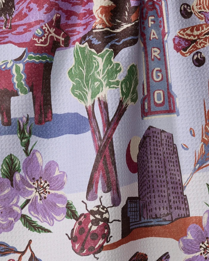 The Geometry "Greetings From North Dakota" dish towel features an illustrated pattern with rhubarb stalks, a Fargo sign, a tall building, a ladybug, purple flowers, a colorful cow, and other local motifs on a textured background.