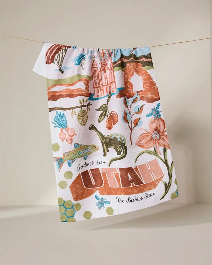 The Geometry Greetings From Utah tea towel features Utah-inspired illustrations—mountains, birds, cherries, and the Salt Lake Temple—with “Greetings from UTAH The Beehive State,” made from recycled materials.