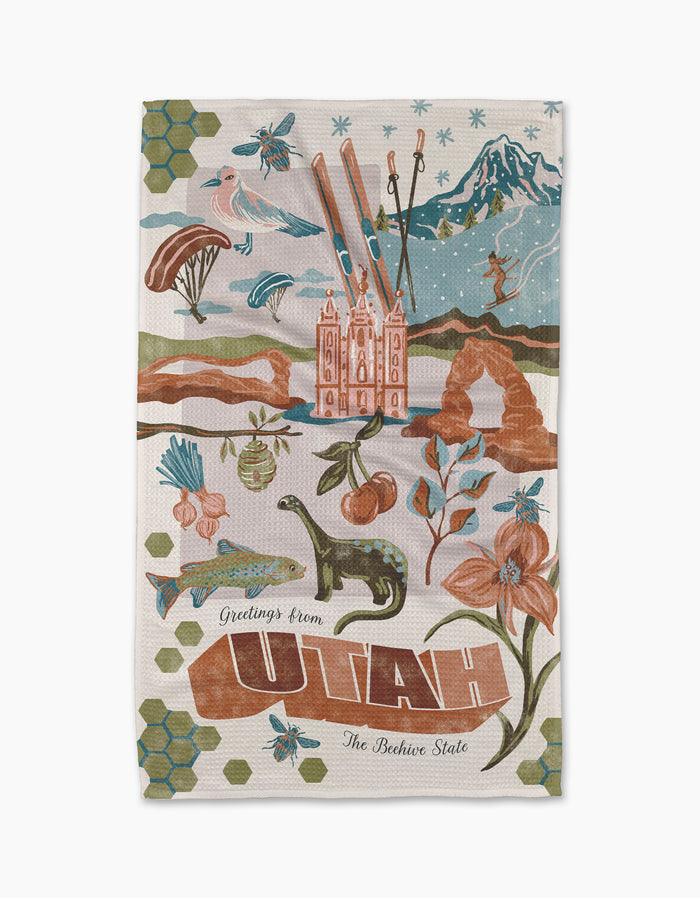 Geometry’s “Greetings From Utah” tea towel features colorful illustrations of Utah landmarks—mountains, Delicate Arch, Salt Lake Temple, dinosaurs, flowers, bees, skiing, and a bison—on quick-drying recycled material.