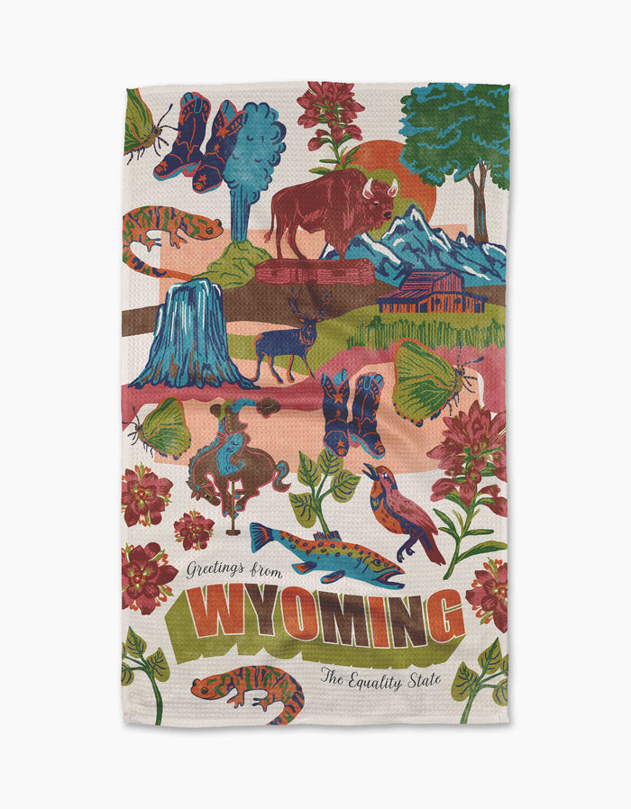 The Geometry "Greetings From Wyoming" kitchen towel showcases colorful illustrations of Wyoming wildlife, mountains, a rodeo rider, flowers, and bold “Greetings from WYOMING The Equality State” lettering at the bottom.
