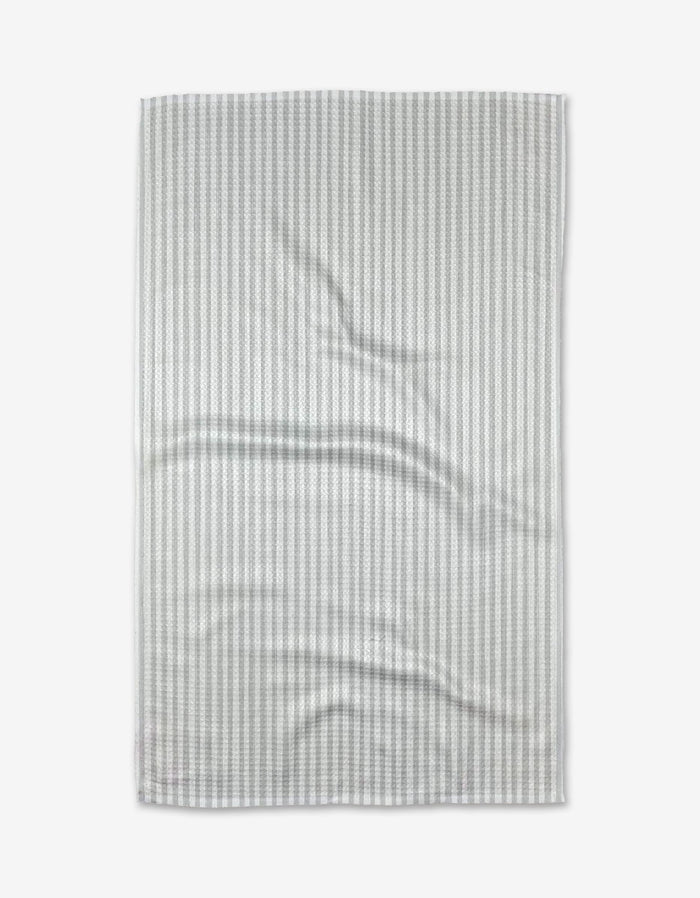 A Grey Salt tea towel by Kailee Wright is laid flat on a white background, showing light gray and white stripes with gentle folds across its ultra-absorbent Geoweave™ fabric.
