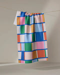 The "Grid" kitchen towel by Jennifer Bouron, made from recycled materials, features a vibrant geometric pattern of blue, pink, green, orange, and yellow rectangles and squares displayed against a neutral background.