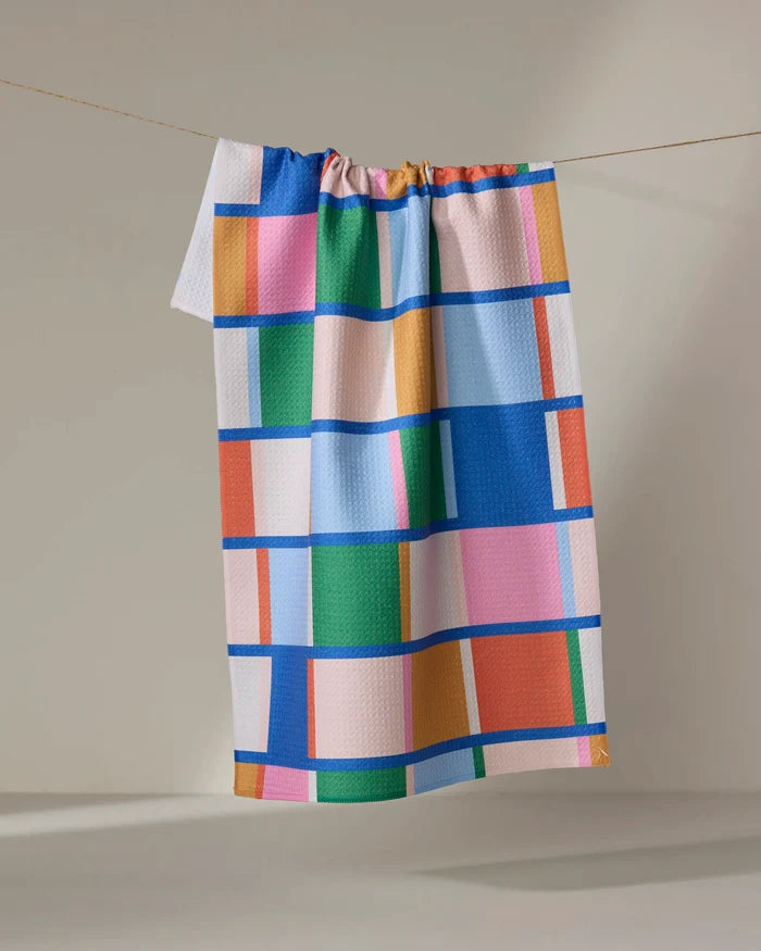 The "Grid" kitchen towel by Jennifer Bouron, made from recycled materials, features a vibrant geometric pattern of blue, pink, green, orange, and yellow rectangles and squares displayed against a neutral background.
