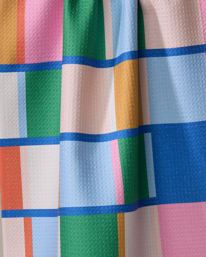 A close-up of Jennifer Bouron's Grid kitchen towel shows a colorful geometric pattern with blue, green, pink, white, orange, and yellow rectangles divided by blue lines on a slightly textured recycled fabric.
