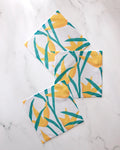 Three GEOMETRY "Growth" dishtowels with yellow flower and green leaf designs are arranged in a geometric, slightly overlapping layout on a white marble surface. This super-absorbent set brightens any kitchen.
